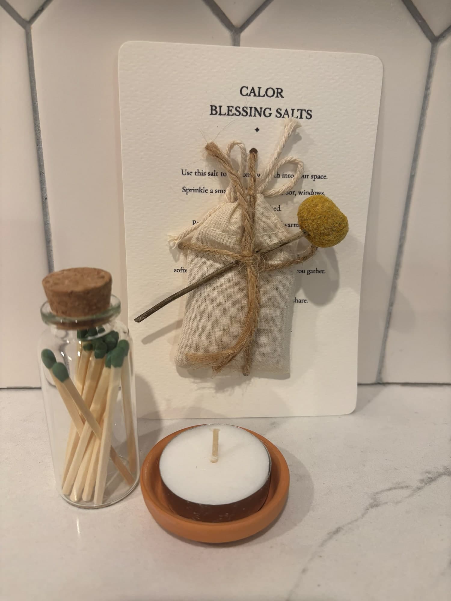 A Calor blessing salts card tied with twine and a dried flower, beside a jar of matches and a tealight in a small terracotta dish - a card and candle gift