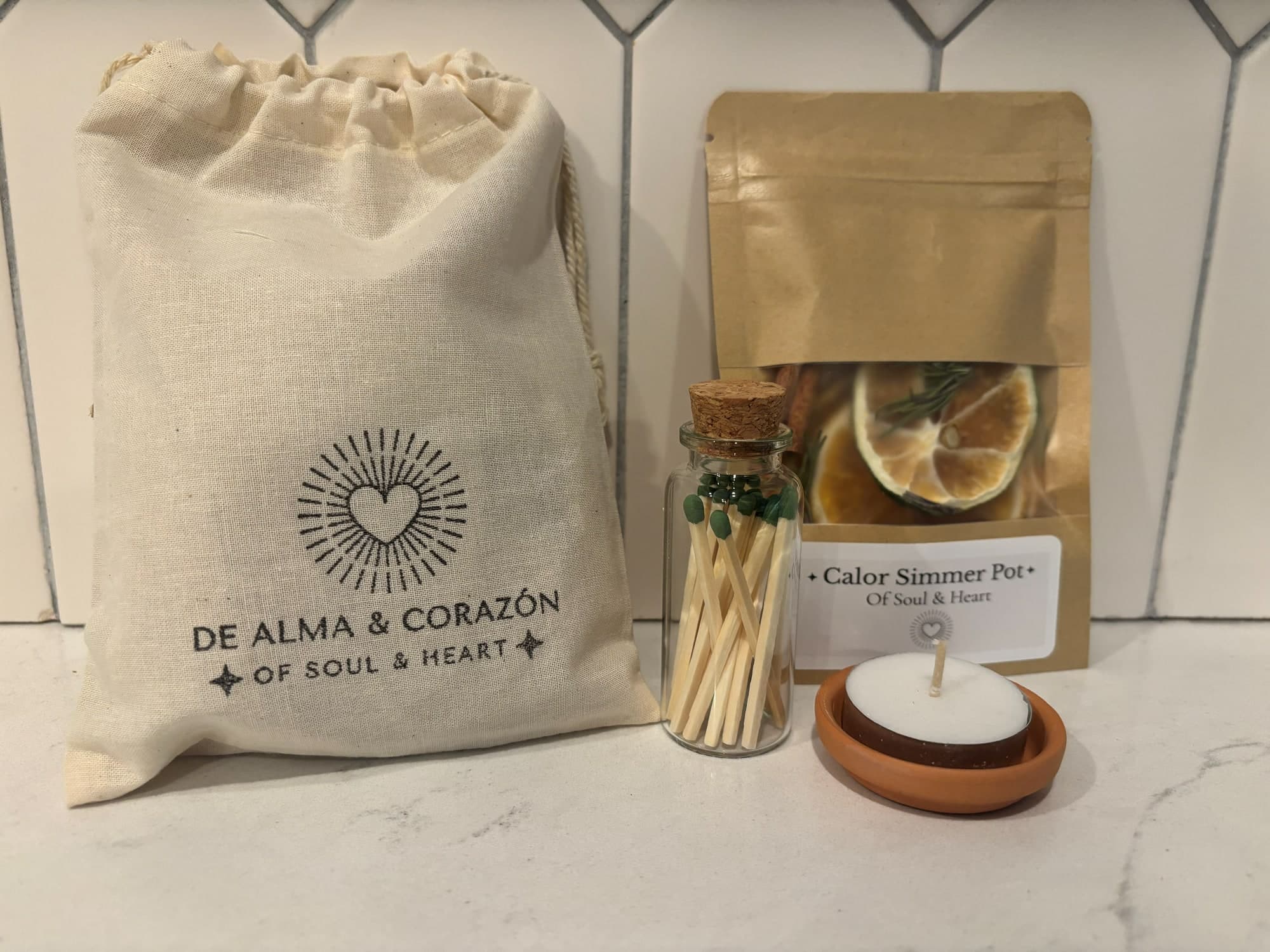 A Calor simmer pot of dried citrus beside a cotton De Alma y Corazón bag, a jar of matches and a tealight in terracotta
