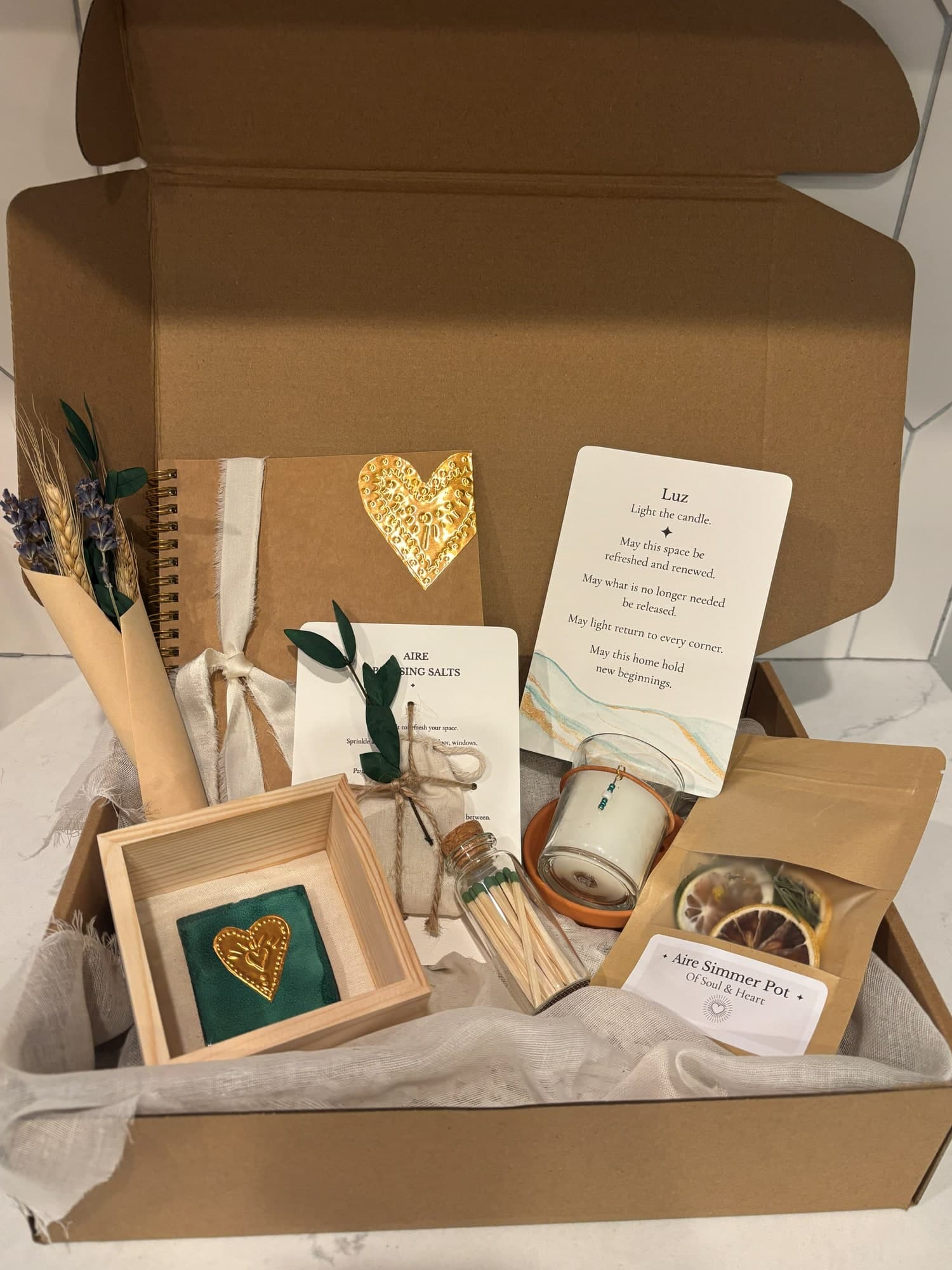 An open Casa gift box with a gold-heart journal, a Luz blessing card, a framed teal tile with a gold heart, a small candle and a simmer pot